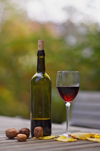 Bottiglia di vino rosso italiano, con accanto un calice mezzo pieno. Sullo sfondo una vigna e sul tavolo alcune noci