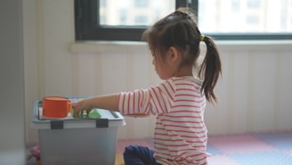 A young child with a ponytail is sitting on a foam mat, engaging with toys placed on a storage box. The child is wearing a pink and white striped shirt and is focused on the activity. Soft light enters through a window in the background.