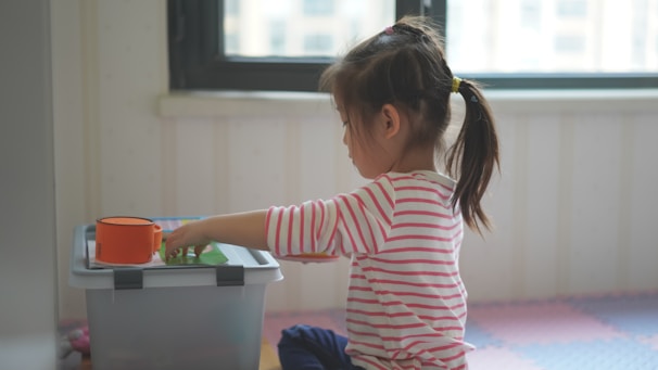 A young child with a ponytail is sitting on a foam mat, engaging with toys placed on a storage box. The child is wearing a pink and white striped shirt and is focused on the activity. Soft light enters through a window in the background.