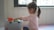 A young child with a ponytail is sitting on a foam mat, engaging with toys placed on a storage box. The child is wearing a pink and white striped shirt and is focused on the activity. Soft light enters through a window in the background.