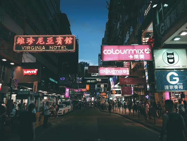 A city street at dusk glowing with orange and blue neon signs reflecting on wet pavement.