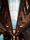 A narrow indoor gallery with a glass ceiling and ornate architecture. The passageway is lined with shops, some displaying various colorful items. There are Turkish flags hanging on either side, and rows of lamps provide warm lighting.