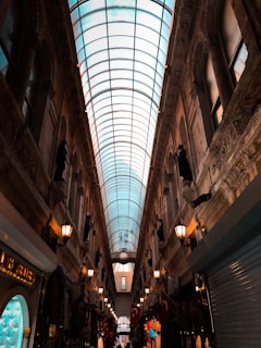 A narrow indoor gallery with a glass ceiling and ornate architecture. The passageway is lined with shops, some displaying various colorful items. There are Turkish flags hanging on either side, and rows of lamps provide warm lighting.