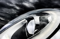 A close-up of a modern car headlight is set against a dark, cloudy sky. The headlight features a sleek, aerodynamic design with LED light strips and reflective surfaces.