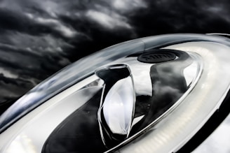 A close-up of a modern car headlight is set against a dark, cloudy sky. The headlight features a sleek, aerodynamic design with LED light strips and reflective surfaces.