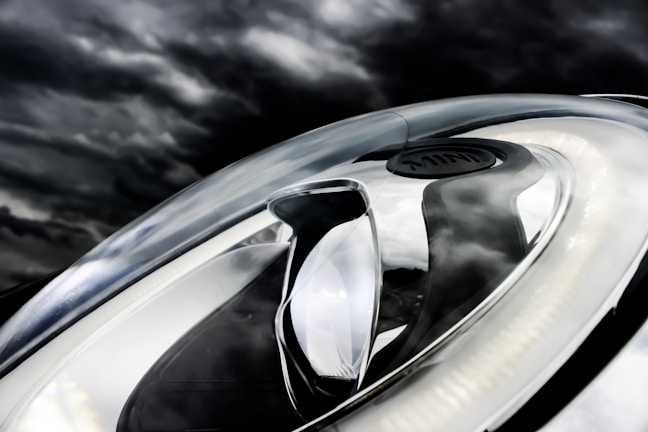 A close-up of a modern car headlight is set against a dark, cloudy sky. The headlight features a sleek, aerodynamic design with LED light strips and reflective surfaces.