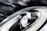 A close-up of a modern car headlight is set against a dark, cloudy sky. The headlight features a sleek, aerodynamic design with LED light strips and reflective surfaces.
