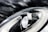A close-up of a modern car headlight is set against a dark, cloudy sky. The headlight features a sleek, aerodynamic design with LED light strips and reflective surfaces.