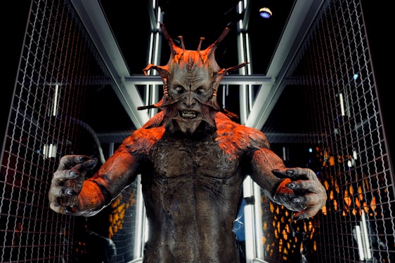 A menacing creature with multiple protrusions from its head stands in a futuristic corridor lined with metallic and glass elements. The creature's skin is textured and appears to have a reddish-brown tone, with sharp, claw-like fingers extended forward. The setting suggests a scene from a science fiction or horror genre.