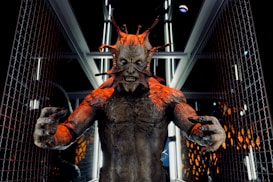 A menacing creature with multiple protrusions from its head stands in a futuristic corridor lined with metallic and glass elements. The creature's skin is textured and appears to have a reddish-brown tone, with sharp, claw-like fingers extended forward. The setting suggests a scene from a science fiction or horror genre.