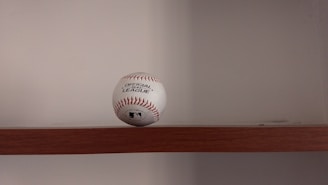 A baseball is placed on a wooden shelf against a plain wall. The baseball features red stitching and has printed labels indicating it is an official league ball.