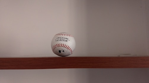 A baseball is placed on a wooden shelf against a plain wall. The baseball features red stitching and has printed labels indicating it is an official league ball.
