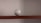A baseball is placed on a wooden shelf against a plain wall. The baseball features red stitching and has printed labels indicating it is an official league ball.