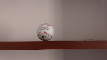 A baseball is placed on a wooden shelf against a plain wall. The baseball features red stitching and has printed labels indicating it is an official league ball.