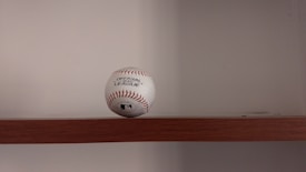 A baseball is placed on a wooden shelf against a plain wall. The baseball features red stitching and has printed labels indicating it is an official league ball.