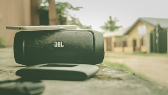 A black JBL portable speaker is placed on a concrete surface with a smartphone resting on top of it. The background includes blurred houses and greenery, creating a casual outdoor setting.