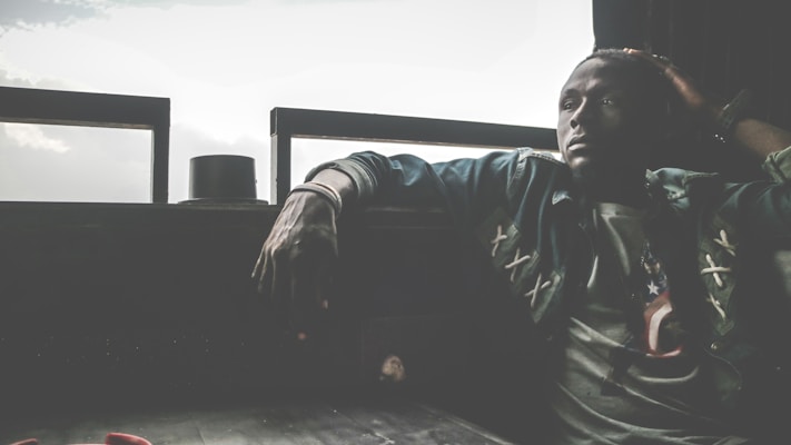 A person sits indoors with a contemplative expression, resting an arm on the back of the seat. The environment is dimly lit with light coming through large windows in the background, creating a contrast. The person appears relaxed yet introspective.