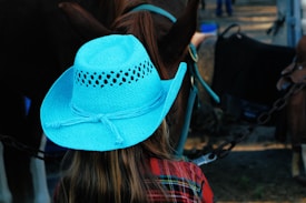 A person with long hair wearing a bright blue cowboy hat stands in front of a horse. The horse's dark brown coat contrasts with the vibrant color of the hat. The person is dressed in a red plaid shirt. The setting appears to be outdoors, possibly at a farm or ranch.