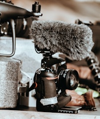 A professional camera equipped with a large, furry microphone is positioned on a surface. Surrounding items include components of a drone and some indistinct background elements, adding a tech-focused atmosphere.