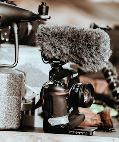 A professional camera equipped with a large, furry microphone is positioned on a surface. Surrounding items include components of a drone and some indistinct background elements, adding a tech-focused atmosphere.