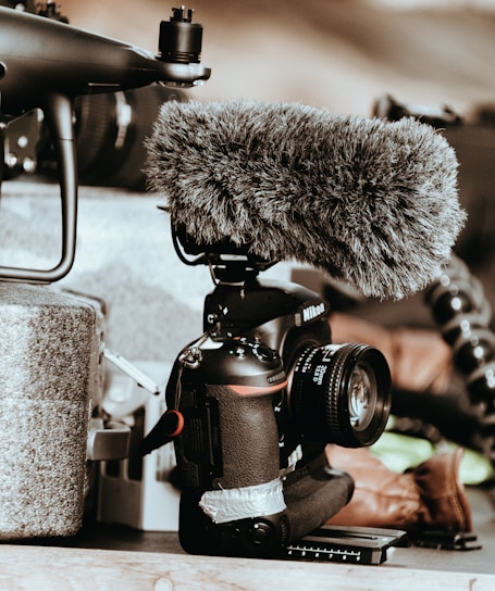A professional camera equipped with a large, furry microphone is positioned on a surface. Surrounding items include components of a drone and some indistinct background elements, adding a tech-focused atmosphere.