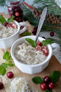 Two white bowls filled with oatmeal garnished with cranberries, shredded coconut, and mint leaves are placed on a wooden surface. A spoon with a decorative handle rests inside one of the bowls. In the background, there is a jar filled with cranberries and a pinecone with pine needles, adding a festive touch.