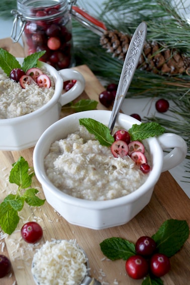 Two white bowls filled with oatmeal garnished with cranberries, shredded coconut, and mint leaves are placed on a wooden surface. A spoon with a decorative handle rests inside one of the bowls. In the background, there is a jar filled with cranberries and a pinecone with pine needles, adding a festive touch.