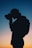 A silhouette of a person with a ponytail is holding a camera in front of their face, set against a vibrant twilight sky with gradient hues transitioning from blue to pink and orange. The person appears to be focusing on capturing an image, and a backpack is visible on their back.