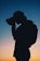 A silhouette of a person with a ponytail is holding a camera in front of their face, set against a vibrant twilight sky with gradient hues transitioning from blue to pink and orange. The person appears to be focusing on capturing an image, and a backpack is visible on their back.