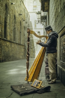 A street musician plays a harp in a narrow, old European alleyway. The musician is wearing a hat and warm clothing, suggesting a chilly day. The harp is intricately carved and stands out against the stone walls of the historic buildings. In front of the musician, there is a cloth spread on the ground with scattered coins, indicating he is performing for tips. The setting has a timeless, rustic charm with ornate street lamps and arched windows visible in the background.