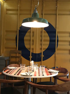 A round table is set with plates, cutlery, napkins, and glasses, beneath a green overhead lamp. The table has a striped pattern on the surface, surrounded by wooden chairs. The background features a yellow wall with vertical support beams and a large blue circle design.