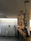 A modern interior space features a long counter with adjustable industrial-style pendant lights hanging from the ceiling. The background includes a wall with vertical rope decorations and tall white planters housing small plants, adding a touch of greenery to the scene. A mix of metal, glass, and minimalist design elements contributes to a contemporary aesthetic.