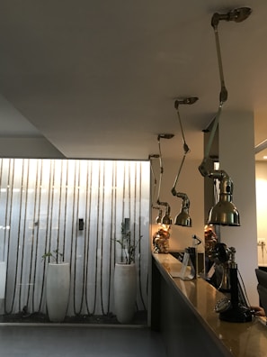 A modern interior space features a long counter with adjustable industrial-style pendant lights hanging from the ceiling. The background includes a wall with vertical rope decorations and tall white planters housing small plants, adding a touch of greenery to the scene. A mix of metal, glass, and minimalist design elements contributes to a contemporary aesthetic.