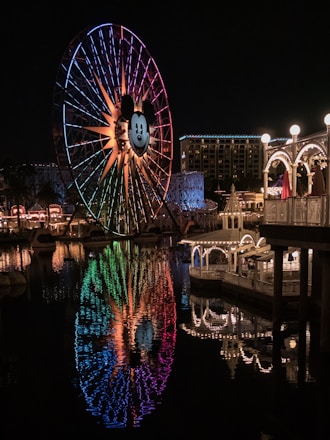 A large Ferris wheel with a prominent cartoon mouse face at its center is illuminated with colorful lights, reflecting beautifully on the water below. The scene is set at night, adding a magical atmosphere. In the background, brightly lit structures and a hotel can be seen.