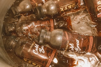 Bottles of cold brew coffee are submerged in ice water. The bottles have labels indicating they are handcrafted with no food coloring. The lids are black, and the liquid inside is dark brown.