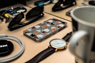 A collection of everyday items laid out on a wooden surface. A smartphone with a cartoon beverage case design is centrally placed, surrounded by sunglasses, a wristwatch, a mug, a coiled cable, keys, and a camera lens cap. The arrangement appears casual yet organized, conveying a sense of preparation or readiness.