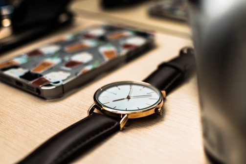 A close-up of a wristwatch with a black leather strap and a golden case is placed on a wooden surface. In the background, a smartphone with a colorful case featuring various beverage illustrations is visible, along with blurred elements that suggest a desk or tabletop environment.