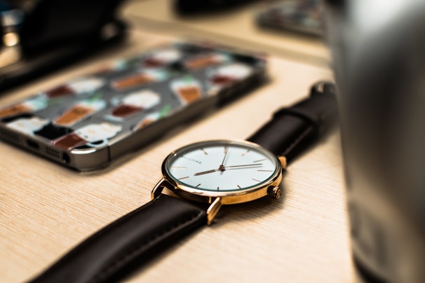 A close-up of a wristwatch with a black leather strap and a golden case is placed on a wooden surface. In the background, a smartphone with a colorful case featuring various beverage illustrations is visible, along with blurred elements that suggest a desk or tabletop environment.
