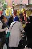 A market scene with several people gathered around a stall. Bunches of grapes in red and yellow hues hang prominently from the overhead structure. The people appear engaged in selecting or purchasing produce. Graffiti on a wall is visible in the background, adding a colorful urban touch.
