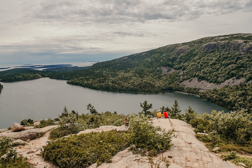 two persons sitting on cliff facing towards body of water