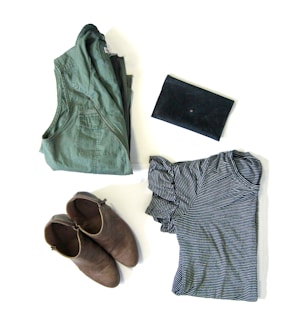 A neatly arranged display of clothing and accessories, including a folded green jacket, a pair of brown ankle boots, a black clutch, and a striped black and white shirt.