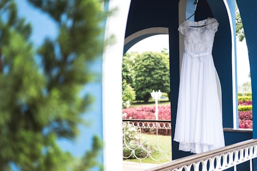 A white wedding dress is hanging on a black hanger inside a blue arched structure. The dress features lace detailing and a ribbon at the waist. In the background, a lush garden with green trees and red-leafed bushes can be seen, along with a white lamp post.