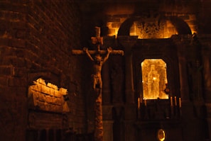 A dimly lit church interior featuring a large crucifix and statues. The stone walls and archways create a somber, reverent atmosphere. Candles are visible, providing a warm glow.