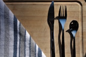 A set of silverware including a knife, fork, and spoon arranged neatly on a wooden cutting board. A striped cloth napkin is partially draped over the corner of the board. The sunlight creates sharp shadows of the utensils, adding contrast to the composition.