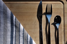 A set of silverware including a knife, fork, and spoon arranged neatly on a wooden cutting board. A striped cloth napkin is partially draped over the corner of the board. The sunlight creates sharp shadows of the utensils, adding contrast to the composition.