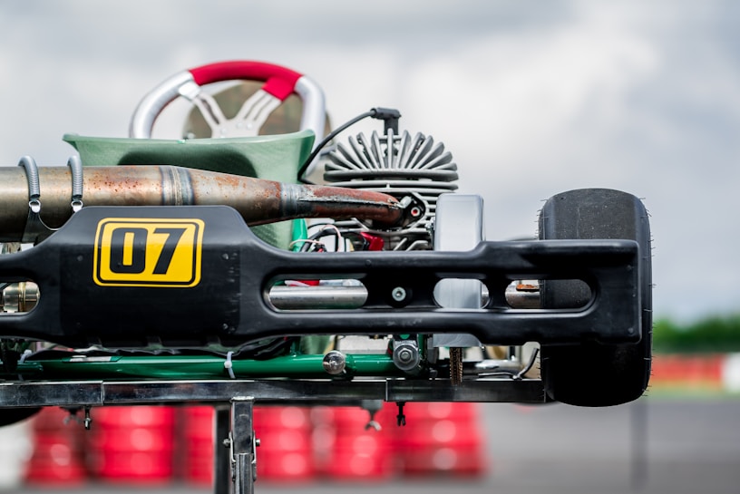 A close-up of a go-kart's rear section, highlighting the exhaust pipe, engine, and a large tire. The number 07 is prominently displayed on the side. The background includes a series of blurred red and white barriers, indicating a racetrack environment.