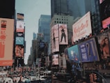 A bustling urban scene filled with large, colorful billboards and advertisements lining the skyscrapers. The streets are crowded with pedestrians and vehicles, creating a lively atmosphere. Buildings are adorned with a variety of ads promoting fashion, music, and technology, capturing the essence of a vibrant commercial district.