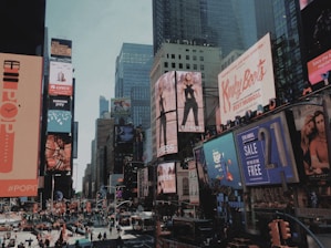 A bustling urban scene filled with large, colorful billboards and advertisements lining the skyscrapers. The streets are crowded with pedestrians and vehicles, creating a lively atmosphere. Buildings are adorned with a variety of ads promoting fashion, music, and technology, capturing the essence of a vibrant commercial district.