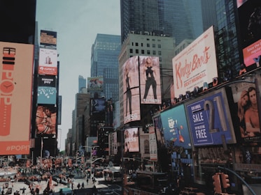 A bustling urban scene filled with large, colorful billboards and advertisements lining the skyscrapers. The streets are crowded with pedestrians and vehicles, creating a lively atmosphere. Buildings are adorned with a variety of ads promoting fashion, music, and technology, capturing the essence of a vibrant commercial district.