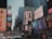 A bustling urban scene filled with large, colorful billboards and advertisements lining the skyscrapers. The streets are crowded with pedestrians and vehicles, creating a lively atmosphere. Buildings are adorned with a variety of ads promoting fashion, music, and technology, capturing the essence of a vibrant commercial district.